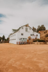 A large white barn with green trim and doors stands on a dirt lot, surrounded by rocks, trees, and a partly cloudy sky.