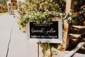 A sign decorated with greenery reads, “Welcome family and friends to the wedding of Hannah + Julio, October 11, 2024, Bailey, CO, Elevation 7,710.”.