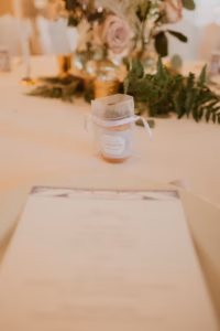 A small jar with a tag and ribbon sits on a decorated table, with flowers and greenery in the background and a blurred menu in the foreground.