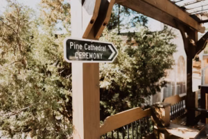 A wooden post with a sign reading "Pine Cathedral Ceremony" and an arrow pointing right, set outdoors with trees and wooden beams in the background.