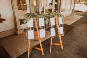 Two wooden easels hold wire grids displaying seating cards with greenery on top, positioned near the entrance of an event venue.