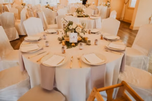 A round table set for a formal event with white tablecloth, covered chairs, plates, menus, cutlery, small jars, and a floral centerpiece in a banquet hall.