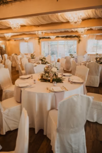 Round tables covered in white cloths with elegant floral centerpieces and neatly arranged place settings are set up in a decorated banquet hall with chandeliers and draped fabric on the ceiling.