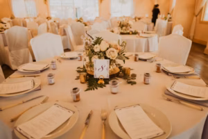 Round banquet table set for an event, decorated with white flowers, greenery, candles, and a table number card, surrounded by white-covered chairs.