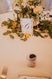 Table centerpiece with greenery, cream flowers, a card displaying the number 6, gold rings, and a small dessert cup in the foreground.