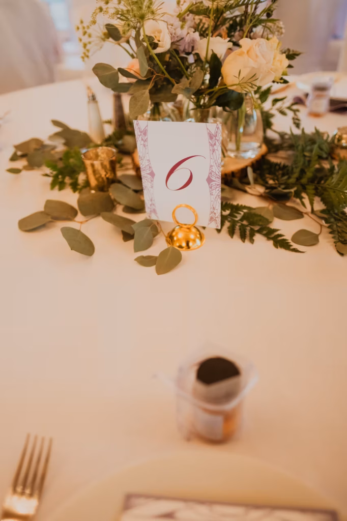 Table centerpiece with greenery, cream flowers, a card displaying the number 6, gold rings, and a small dessert cup in the foreground.