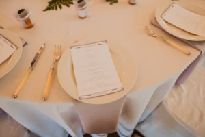 A round table set for a meal with white plates, beige-handled cutlery, menus on each plate, and small favors; decorated with a light tablecloth and green leaves.