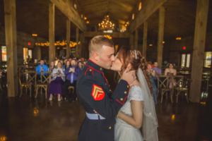 A bride and groom share a kiss at their wedding ceremony in a rustic hall, with guests seated and watching in the background.