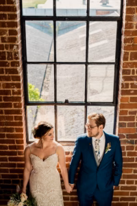 A bride and groom stand holding hands in front of a large window set in a brick wall, looking at each other.