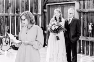 A woman reads from a paper while another woman in a wedding dress holding a bouquet stands with a man in a suit in a rustic wooden venue.