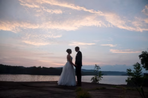 A bride and groom stand facing each other and holding hands by a lakeside at sunset, with trees and calm water in the background.