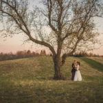 A bride and groom stand together under a large tree in an open grassy field at sunset.