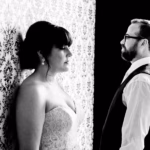 A bride in a strapless dress stands facing a patterned wall while a man in a vest and glasses stands facing her. The image is in black and white.