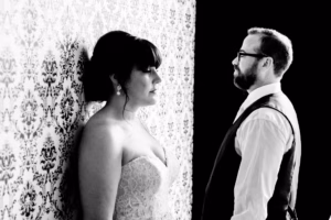 A bride in a strapless dress stands facing a patterned wall while a man in a vest and glasses stands facing her. The image is in black and white.
