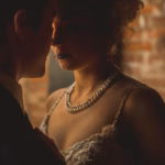 A man in a suit and a woman in a white dress with a pearl necklace stand close together in dim lighting, facing each other.