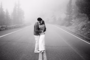 A couple stands kissing in the middle of an empty, misty road surrounded by trees and rocks.
