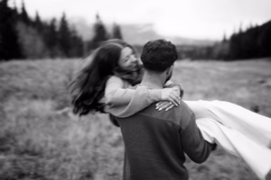 A man carries a smiling woman in his arms through a grassy field with trees in the background. The image is black and white and slightly blurred.