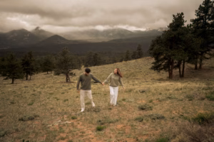 A man and woman holding hands walk up a grassy hill, with pine trees and cloudy mountains in the background.
