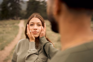 A woman looks directly at the camera while a man, seen from behind, gently holds her face with both hands on a dirt path outdoors.