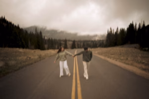 Two people holding hands walk along the center of an empty road surrounded by trees and mountains under a cloudy sky.