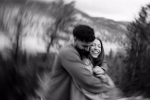 A black and white photo of a couple embracing outdoors, with a blurred background effect and trees in the distance.