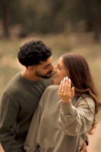 A woman shows an engagement ring on her hand while standing close to a man outdoors; both are dressed in casual clothing.