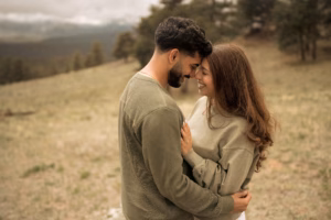 A man and woman stand closely outdoors, touching foreheads and smiling at each other, with trees and hills in the background.