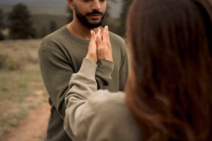 Two people standing outdoors face each other, gently touching palms, both wearing olive green long-sleeve shirts. The background is blurred with trees and grass visible.
