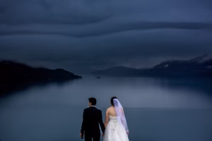 A bride and groom stand hand in hand facing a calm lake under a cloudy, overcast sky, surrounded by mountains.