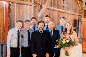 A group of seven men and one woman in formal attire pose indoors; the woman holds a bouquet of flowers and wears a white dress. The background features wooden walls and beams.