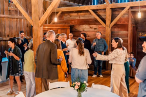 A group of people stand and converse in a rustic wooden room, some holding drinks, with a round table and floral centerpiece in the foreground.