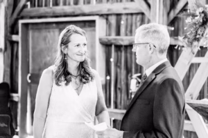 A woman in a white dress stands facing a man in a suit inside a rustic wooden building, engaged in conversation.