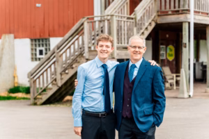 Two men in dress shirts and ties stand side by side, smiling, with one arm around each other's shoulders, in front of a wooden staircase and red building.