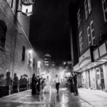 A bride and groom walk down a rainy alley at night, surrounded by people blowing bubbles and taking photos; their shadows are cast on the brick walls.