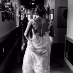A woman in a lace wedding dress walks down a hallway, seen from behind in black and white.