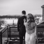 A bride in a white dress approaches a groom in a suit from behind on an outdoor balcony, with a snowy landscape in the background.
