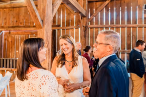Three adults, two women and one man, stand indoors at a rustic wooden venue, smiling and holding drinks while conversing at a social event.