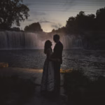 A couple stands facing each other at dusk by a river and waterfall, silhouetted against the fading light.