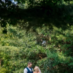 A bride and groom stand holding hands outdoors in front of green trees, looking at each other.
