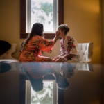 Two women in floral robes sit in a softly lit room; one applies makeup to the other, who is seated near a window, with their reflection visible on a shiny surface below.