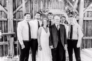 A group of six men and one woman, all dressed formally, pose together inside a wooden barn decorated with string lights and flowers.