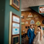 A bride and groom stand together indoors at a bar with brick walls decorated with various brewery signs and vintage decor.