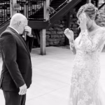 A bride in a lace wedding dress and an older man in a suit wipe away tears while standing outdoors near stairs.