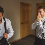 Two men in white dress shirts and suspenders stand indoors; one smiles with hands covering his mouth while the other looks down, holding a garment bag.