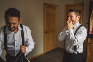 Two men in white dress shirts and suspenders stand indoors; one smiles with hands covering his mouth while the other looks down, holding a garment bag.