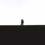 A bride and groom stand closely together on a grassy field with a white sky background, silhouetted against the horizon.