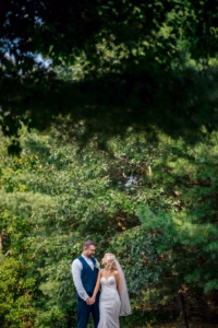 A bride and groom stand outdoors holding hands, surrounded by green trees, with sunlight filtering through the foliage.