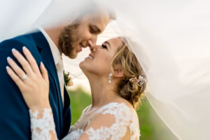 A bride and groom embrace closely, smiling with their noses touching, under a sheer white veil outdoors.