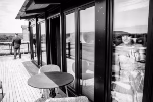 A woman in a wedding dress looks out from behind a glass door, touching the glass, while a man stands on a wooden deck outside with his back turned.