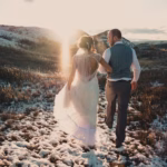 A bride and groom walk together on a snowy path through a field at sunset, with hills in the background.